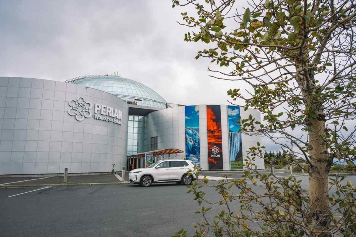 Perlan Museum de Reykjavík Musée Perlan à Reykjavik—dôme argenté et banderoles à l’entrée—avec un parking presque vide et un SUV blanc devant.