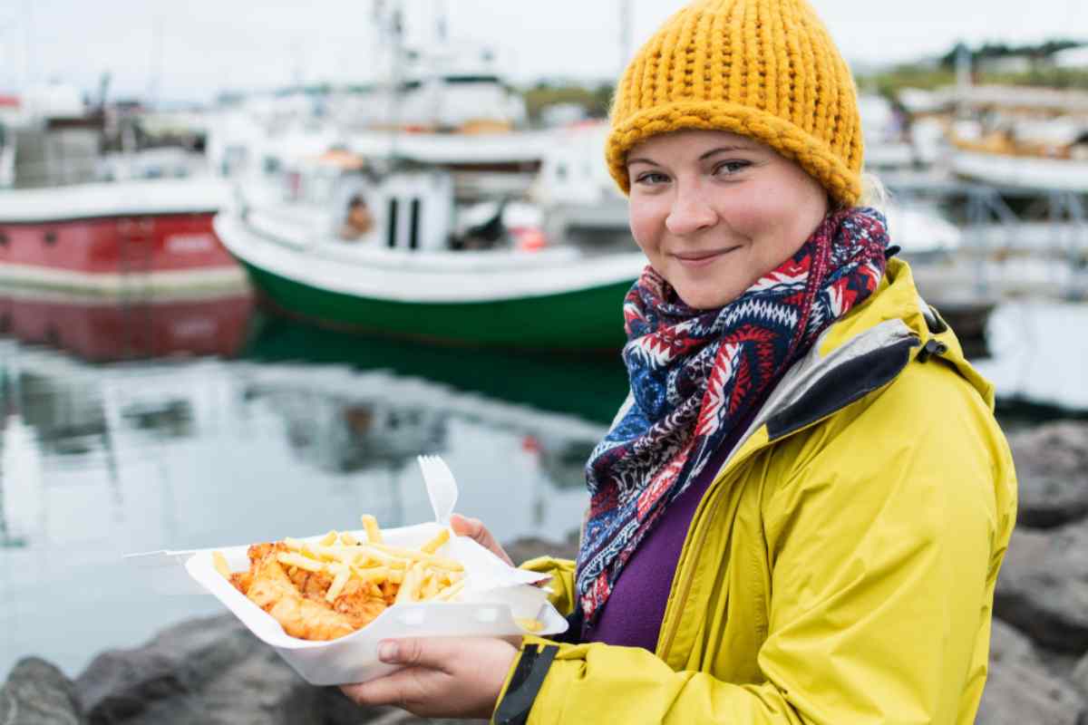 Femme en veste jaune mangeant du fish and chips près d’un port en Islande.