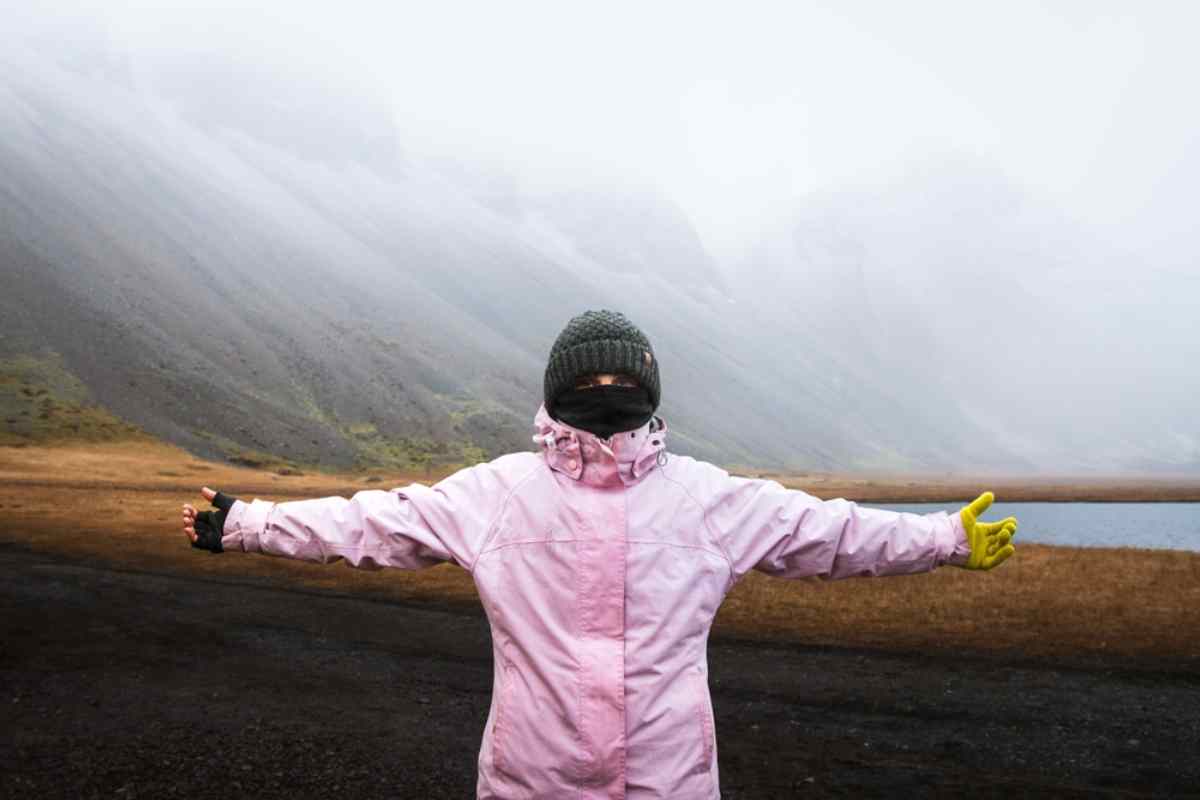 Voyageuse en veste rose et bonnet d’hiver, bras ouverts, dans des montagnes islandaises embrumées.