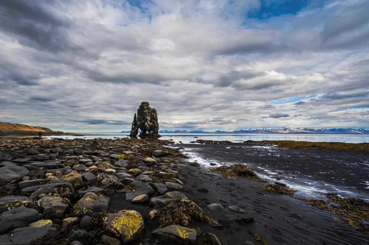 Péninsule de Vatnsnes en Islande Rivage rocheux de sable noir à marée basse avec le stack Hvítserkur sur la péninsule de Vatnsnes, montagnes et ciel nuageux au loin.