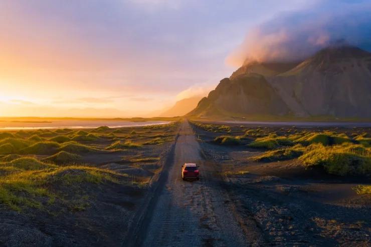 Voiture rouge roulant sur une piste de gravier au coucher du soleil, entre sable noir et dunes vertes, avec des montagnes brumeuses et un ciel lumineu