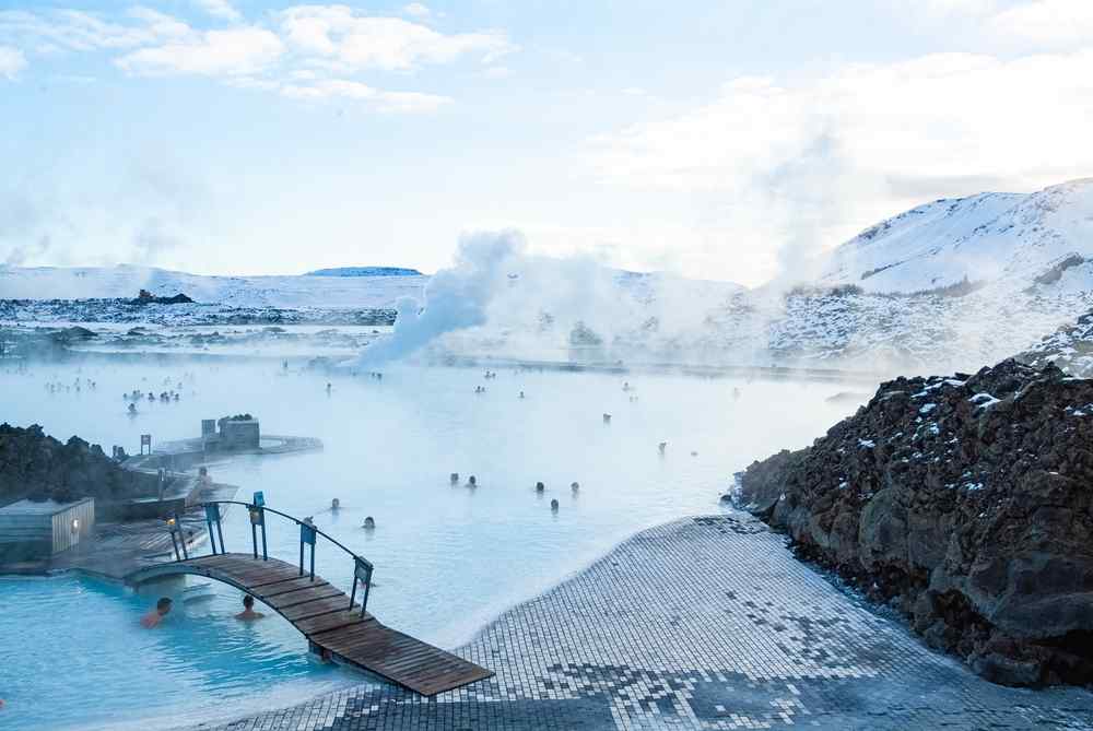 Personnes se baignant dans le spa g&eacute;othermal Blue Lagoon en Islande, entour&eacute;es de vapeur, de roches de lave et de paysages enneig&eacute;s.