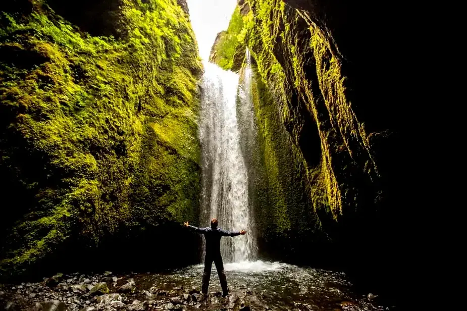 Un randonneur debout, les bras tendus devant la cascade de Nauth&uacute;safoss, entour&eacute; de falaises couvertes de mousse et de verdure luxuriante en Islande. La cascade tombe dans une piscine peu profonde, cr&eacute;ant une sc&egrave;ne sereine et pittoresque.