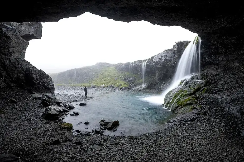 Un randonneur debout pr&egrave;s de la piscine tranquille &agrave; la base de la cascade de Nauth&uacute;safoss, vue de l'int&eacute;rieur d'une formation rocheuse ressemblant &agrave; une grotte. La cascade tombe sur des roches couvertes de mousse dans une piscine claire et peu profonde, entour&eacute;e d'un paysage brumeux et verdoyant en Islande.
