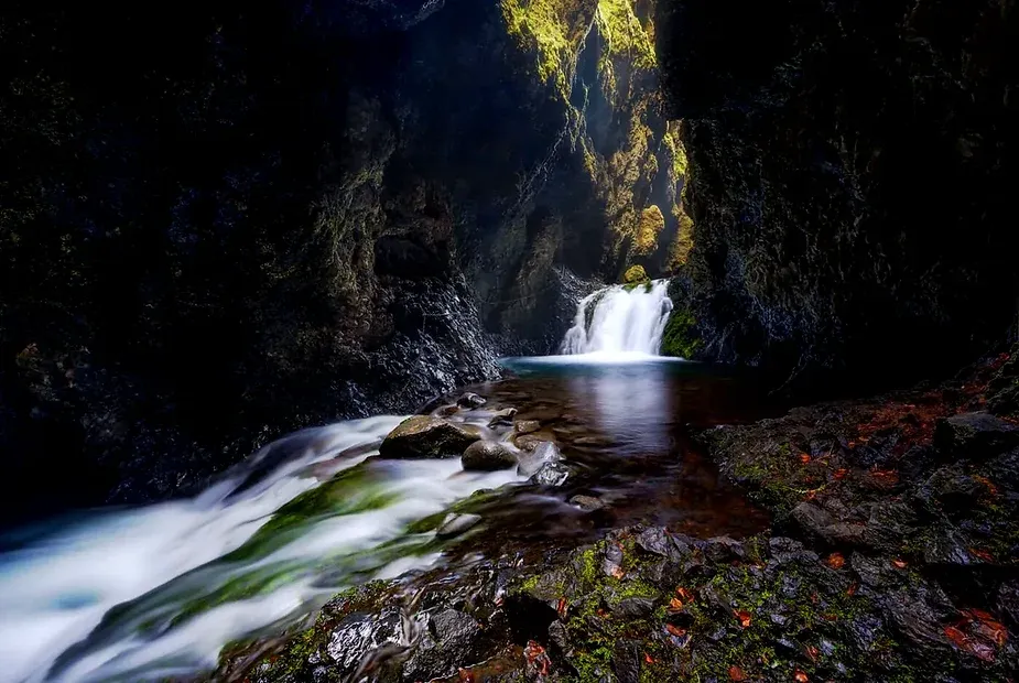 Une vue pittoresque de la cascade de Nauth&uacute;safoss qui se d&eacute;verse dans une piscine sereine au sein d'un gorge rocheux couvert de mousse. La cascade est entour&eacute;e de v&eacute;g&eacute;tation luxuriante et les murs rocheux de la grotte cr&eacute;ent une atmosph&egrave;re dramatique et enchanteresse en Islande.