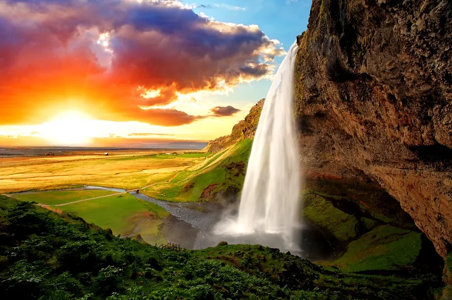 Chute d'eau de Seljalandsfoss en Islande au lever du soleil, avec des couleurs vibrantes illuminant le ciel et les champs verts en dessous.