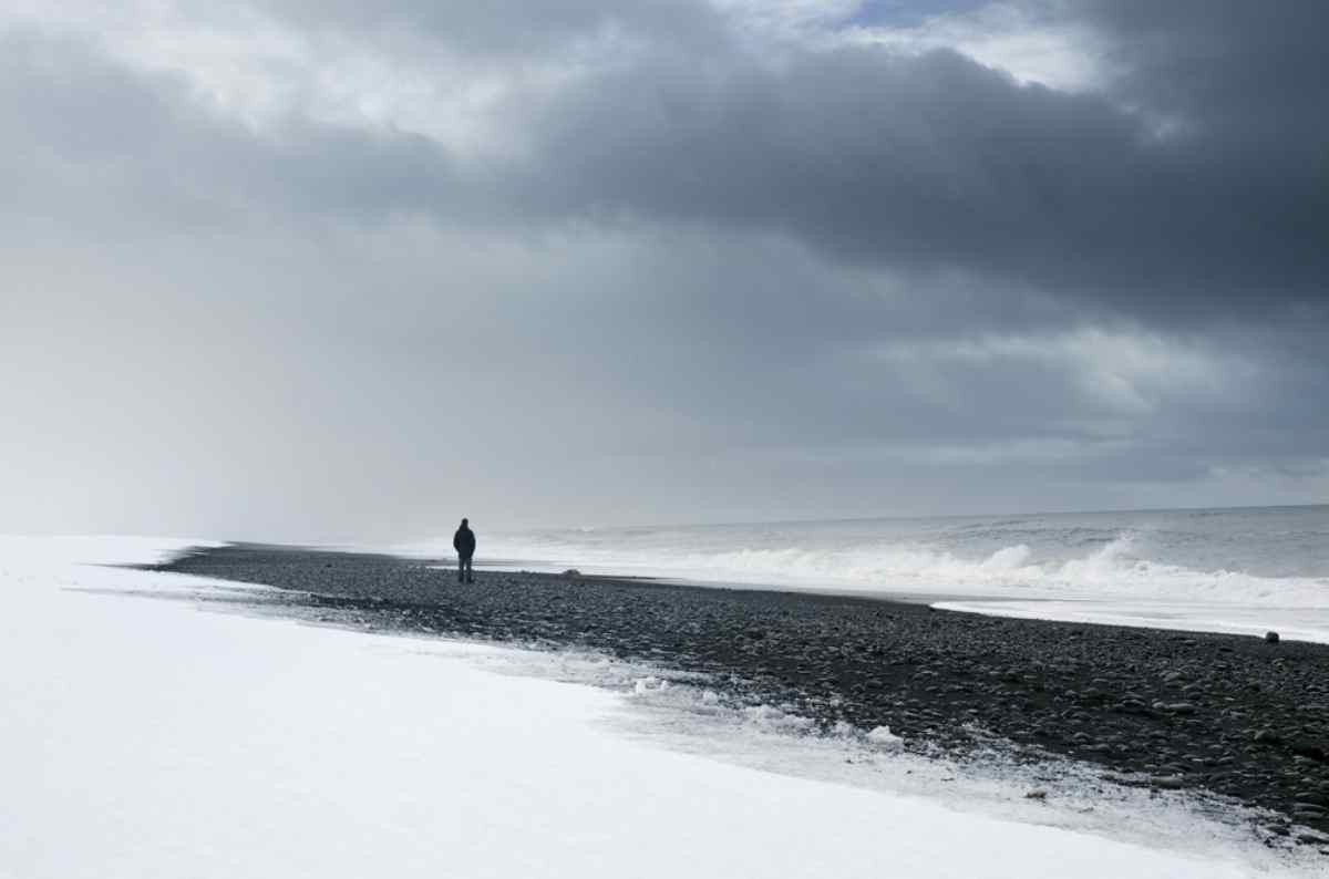 Viaggiatore solitario che cammina lungo una spiaggia di sabbia nera parzialmente coperta di neve in Islanda.