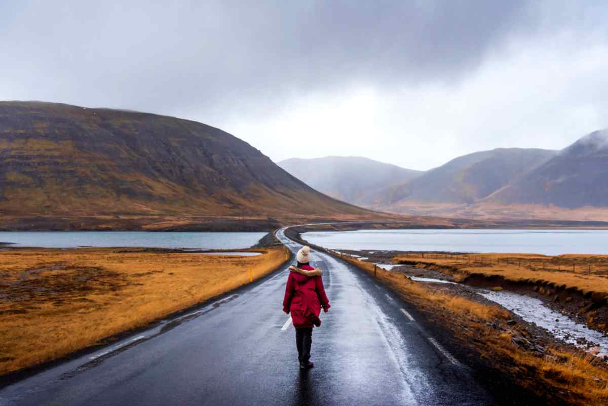 Autunno in Islanda Viaggiatore con parka rosso che cammina su una strada bagnata a due corsie tra laghi e colline autunnali brune in Islanda, con nubi basse e montagne lontane di fronte.