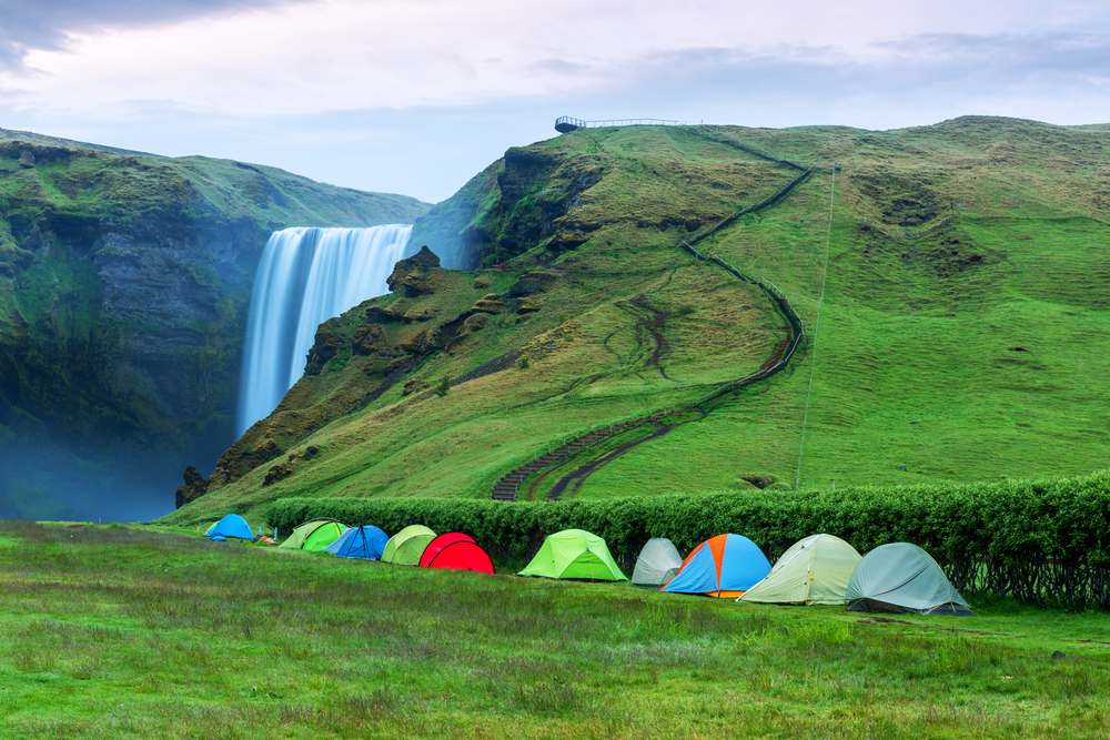 Tende colorate in un campeggio in Islanda, con una grande cascata e colline verdi sullo sfondo.