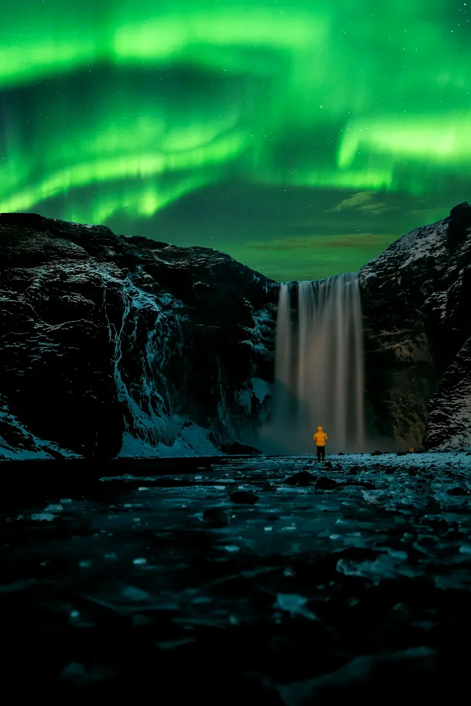 Ammira la cascata Skógafoss sotto le aurore boreali in Islanda: uno spettacolo notturno mozzafiato Una persona con una giacca gialla in piedi di fronte alla cascata Skógafoss illuminata in Islanda, con le aurore boreali che brillano vividamente nel cielo notturno.