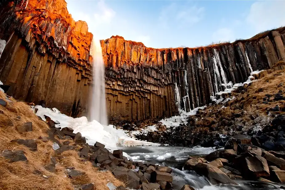 Cascata di Svartifoss che scorre lungo colonne basaltiche scure durante l'inverno in Islanda. Le formazioni rocciose sono parzialmente coperte di ghiaccio e neve, creando un contrasto drammatico. La cascata si riversa in una piccola pozza parzialmente ghiacciata, circondata da terreni accidentati e chiazze di neve.