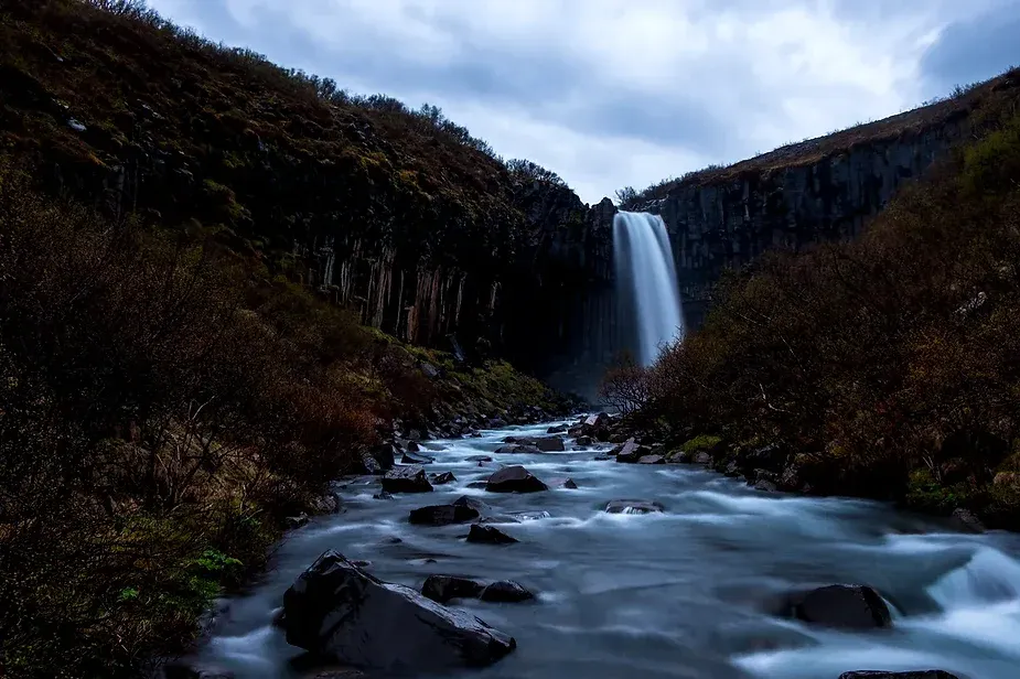 Cascata di Svartifoss illuminata di notte, che scorre lungo colonne basaltiche scure in Islanda. La vegetazione circostante è appena visibile al buio, e la cascata scende in un corso d'acqua roccioso, creando un'atmosfera serena e mistica.
