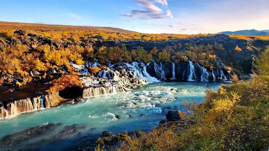 Veduta panoramica della cascata di Hraunfossar in Islanda, con acqua blu limpida che scorre su rocce laviche e circondata da vivaci foglie autunnali s